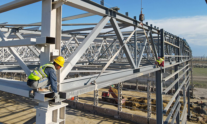 進(jìn)入施工旺季，新疆塔城重點(diǎn)開(kāi)發(fā)開(kāi)放試驗(yàn)區(qū)工程建設(shè)有序進(jìn)行，來(lái)自中鐵十五局等多家單位的工人晝夜奮戰(zhàn)在建設(shè)現(xiàn)場(chǎng)。新疆塔城重點(diǎn)開(kāi)發(fā)開(kāi)放試驗(yàn)區(qū)是我國(guó)西北地區(qū)第一個(gè)沿邊重點(diǎn)開(kāi)發(fā)開(kāi)放試驗(yàn)區(qū)。圖為工人在20余米高的新疆塔城重點(diǎn)開(kāi)發(fā)開(kāi)放試驗(yàn)區(qū)一處風(fēng)機(jī)葉片制造項(xiàng)目頂部進(jìn)行屋面檁條安裝作業(yè)（5月18日無(wú)人機(jī)拍攝）。新華社記者 高晗 攝