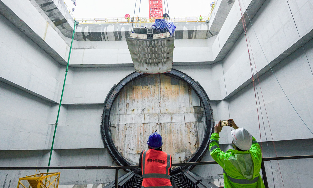 為隧道建設量身定制的盾構機“英雄號”全長134米，開挖直徑14.8米。連日來，盾構機主驅動、刀盤、拼裝機等已運輸下井、完成組裝，后期將進行盾構機管線連接和整機調試工作，即將開始盾構隧道掘進。圖為施工人員在現(xiàn)場緊盯盾體下井定位組裝。